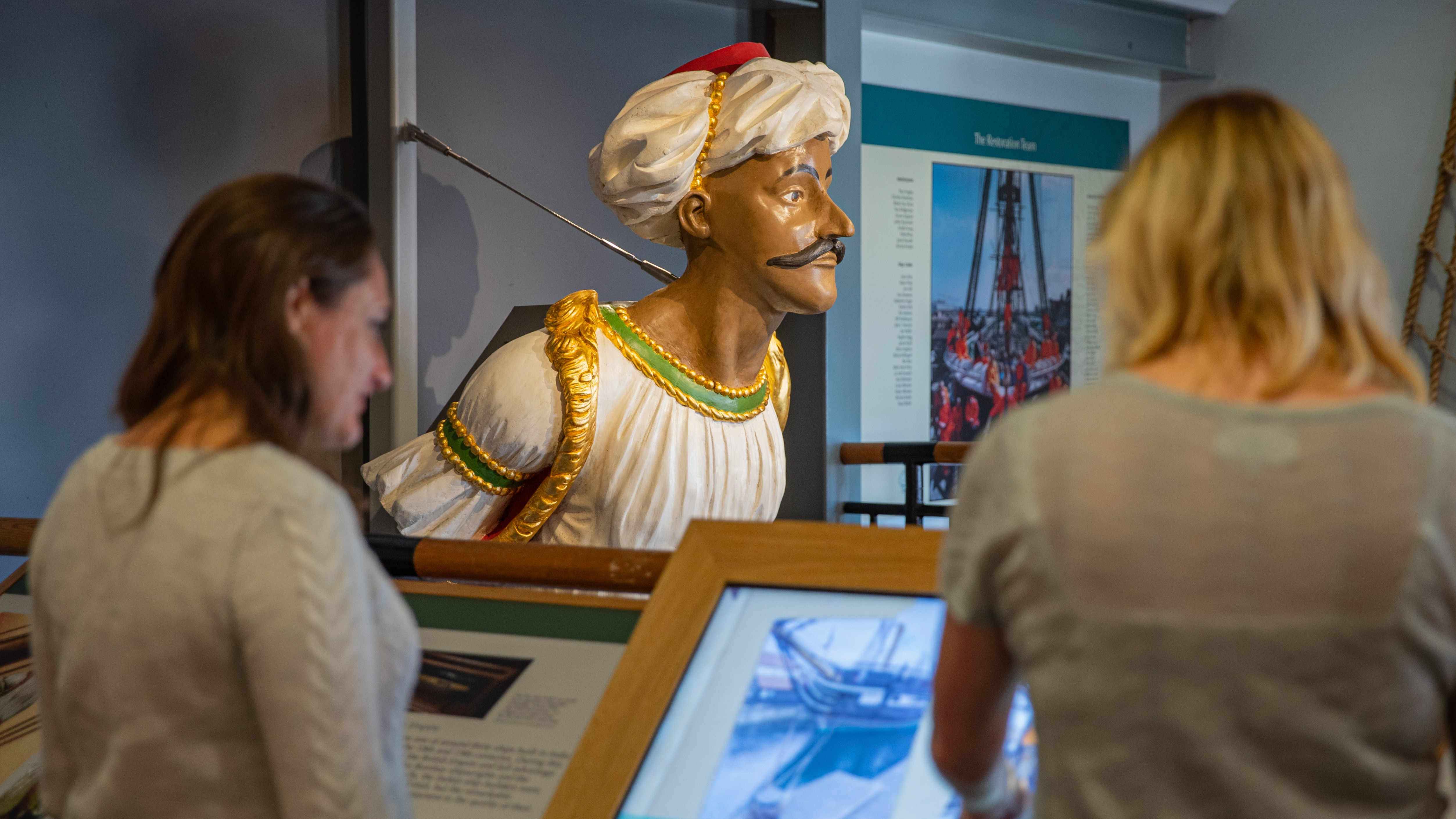 Two visitors look at a painted ship's figurehead depicting a man wearing a white turban and decorated robes in Royal Navy Museums: Hartlepool