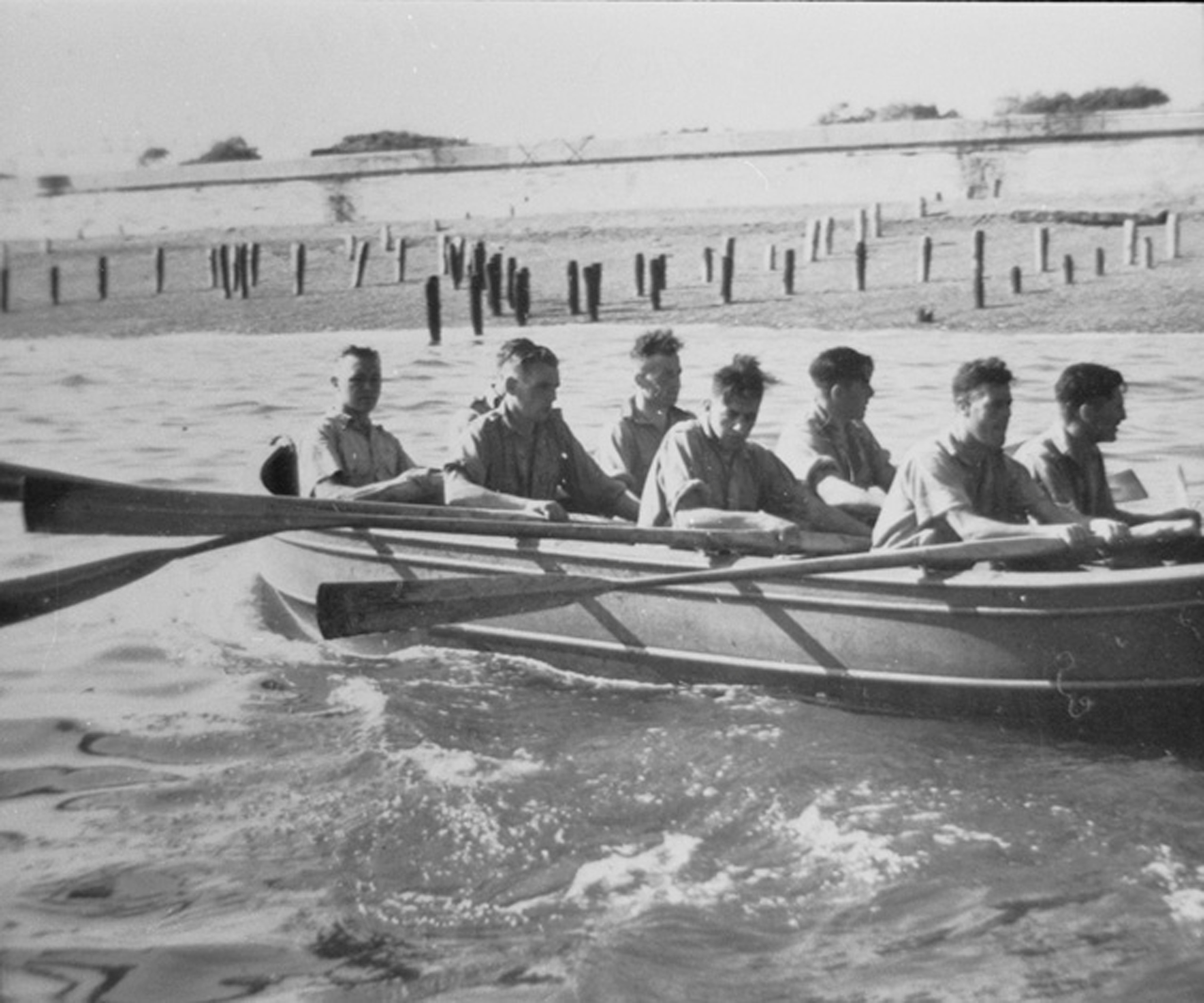 Archive black and white photograph of servicemen rowing a boat near a beach with wooden groynes