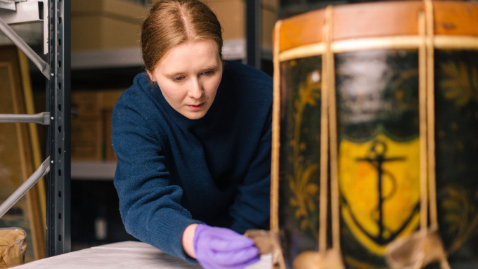 Curator in purple gloves examining a painted band drum with an anchor design, in museum storage