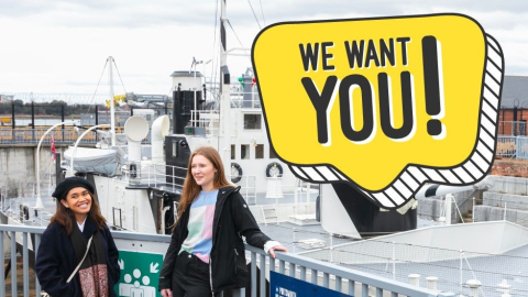 Two teenagers pose in front of HMS M.33 with a yellow speech bubble saying "We want you!".