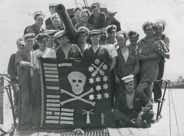 A group of sailors stand behind a handmade Jolly Roger flag. 