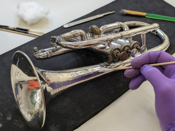 A gloved hand cleans a trombone with a tiny brush. 