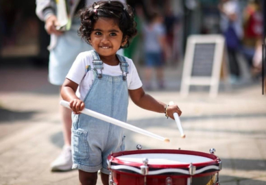 A small child holding drumsticks stood playing a drum.