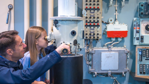 A girl looks through a periscope with her father watching alongside her, in the Search & Rescue exhibit at Royal Navy Museums: Naval Aviation