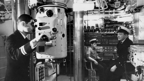 Black and white photograph of the control room of HMS Resolution, a Royal Navy submarine, showing naval officers and a crew member operating equipment including a periscope