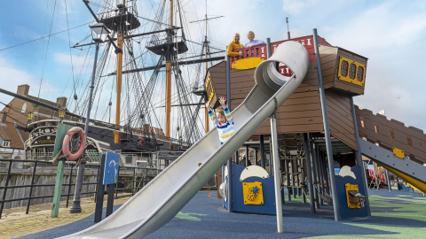A young child slides down the slide of a ship-themed adventure playground with arms raised, with two adults watching from above and HMS Trincomalee visible in the background