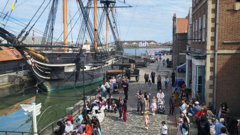 HMS Trincomalee moored at Hartlepool's historic quay, with visitors in period costume walking and gathered along the cobbled dockside in front of brick buildings.