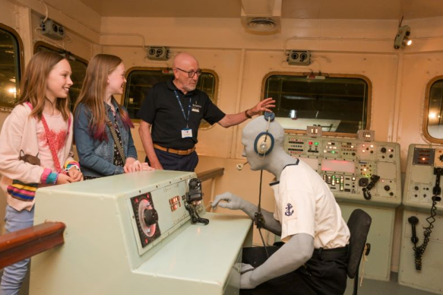 A volunteer show two young girls what the bridge of an aircraft carrier looks like
