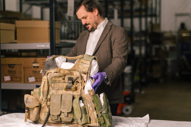 A curator wearing a purple glove examines a camouflage body armour vest, marked 'RY870P', laid out on a table in a museum storage area with shelving in the background