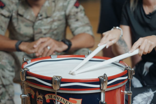Close-up of a visitor being shown how to play a Royal Marines side drum, drumsticks crossed on the drumhead, guided by a hand from someone in military uniform.