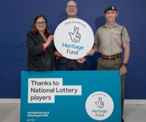 Matthew Sheldon on stage with a National Lottery representative and a Royal Marine. 