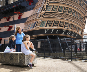 A mother and two children sit outside HMS Victory, a wooden ship. 