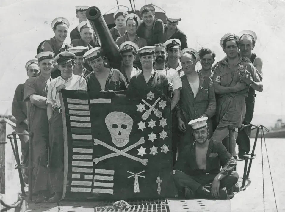 A group of sailors stand behind a handmade Jolly Roger flag. 